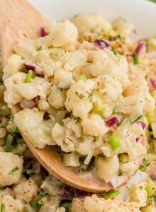 Wooden spoon scooping cauliflower mock potato salad from a large serving bowl of salad. There are some green onions in the background.
