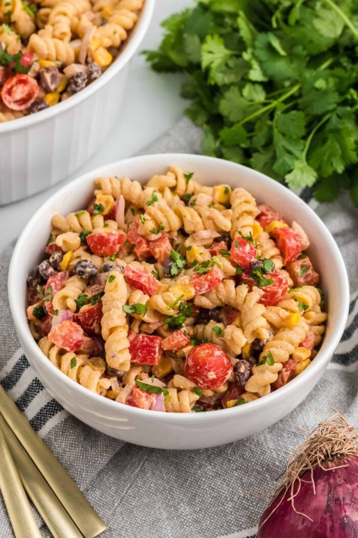 A bowl of creamy vegan Southwestern pasta salad with black beans, peppers and corn. There's cilantro in the background and a larger serving bowl of the salad.