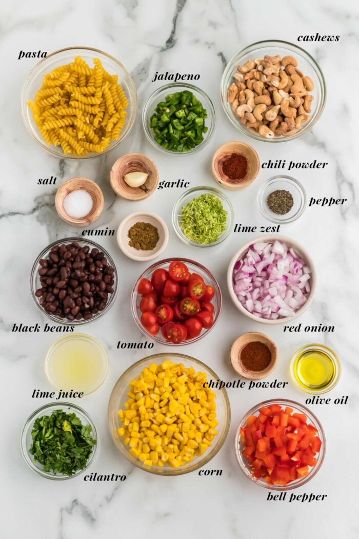 All the ingredients gathered for making a vegan southwest pasta salad with bell pepper, red onion, jalapeno and cashew sauce. Each ingredient is labelled with text.
