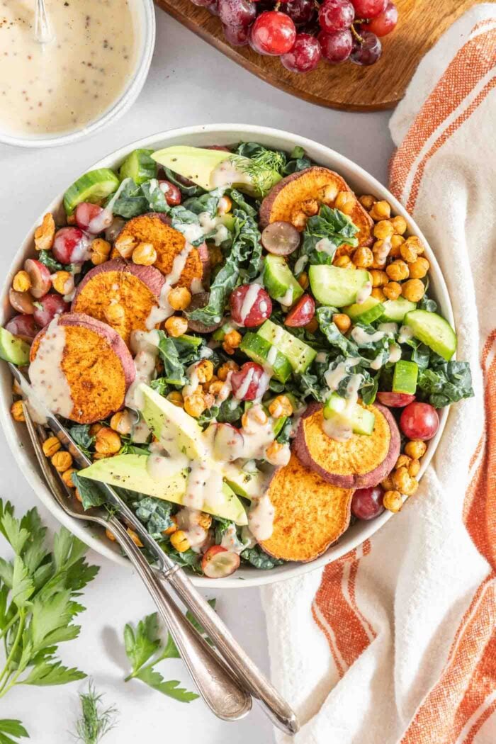 Overhead view of a bowl of Tuscan kale salad with sweet potato, chickpeas, avocado, cucumber, grapes and a creamy dressing drizzled over top.