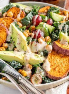 A bowl of tuscan kale salad with avocado, sweet potato, chickpeas, grapes, fresh herbs and a creamy dressing on top. A knife and fork rest on the side of the bowl and there is a bunch of grapes in the bacground.