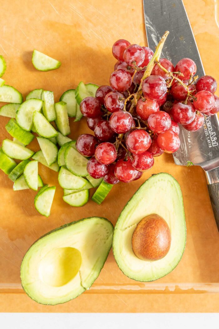 A bunch of red grapes, some sliced cucumber and a halved avocado with the pit in one half on a cutting board with a knife.