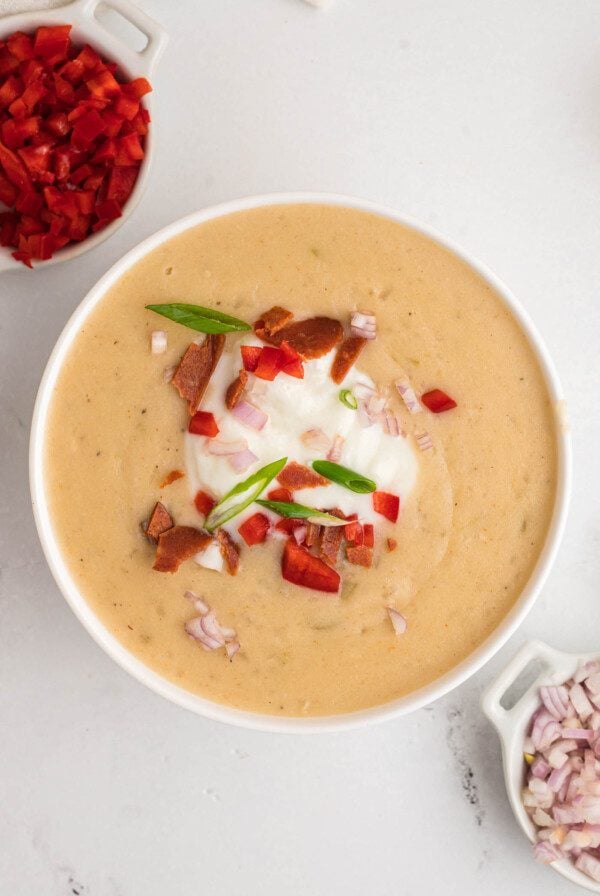 Overhead view of a bowl of creamy potato soup topped with sour cream, bell pepper, bacon, scallions and red onion. There are small dishes of red pepper and red onion beside the bowl.