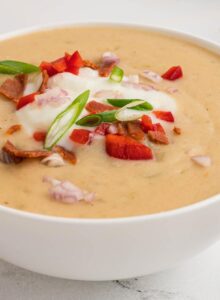 Angled view of a bowl of creamy potato soup topped with sour cream, vegan bacon bits, finely chopped scallions, red onion and bell pepper.