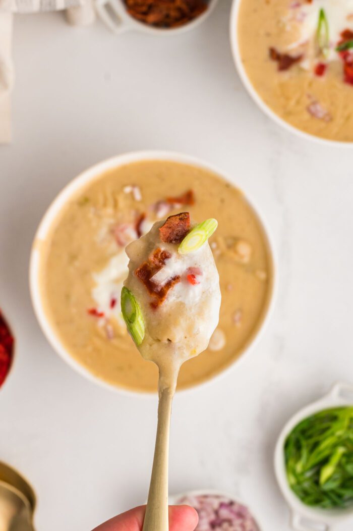 A spoonful of creamy potato soup on a spoon held by a hand. There is some sour cream in the soup and a few bits of green onion and bell pepper.