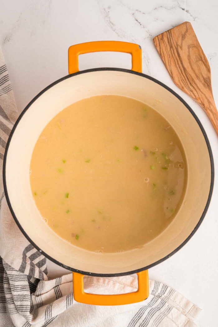 A creamy beige mixture with bits of celery, shallot and garlic in it cooking in a large soup pot with handles. The mixture is the base of a vegan potato soup.