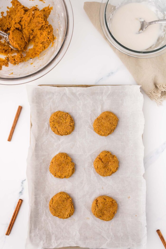 6 baked soft pumpkin cookies on a parchment-paper lined baking tray with a glass mixing bowl of cookie dough beside the tray.