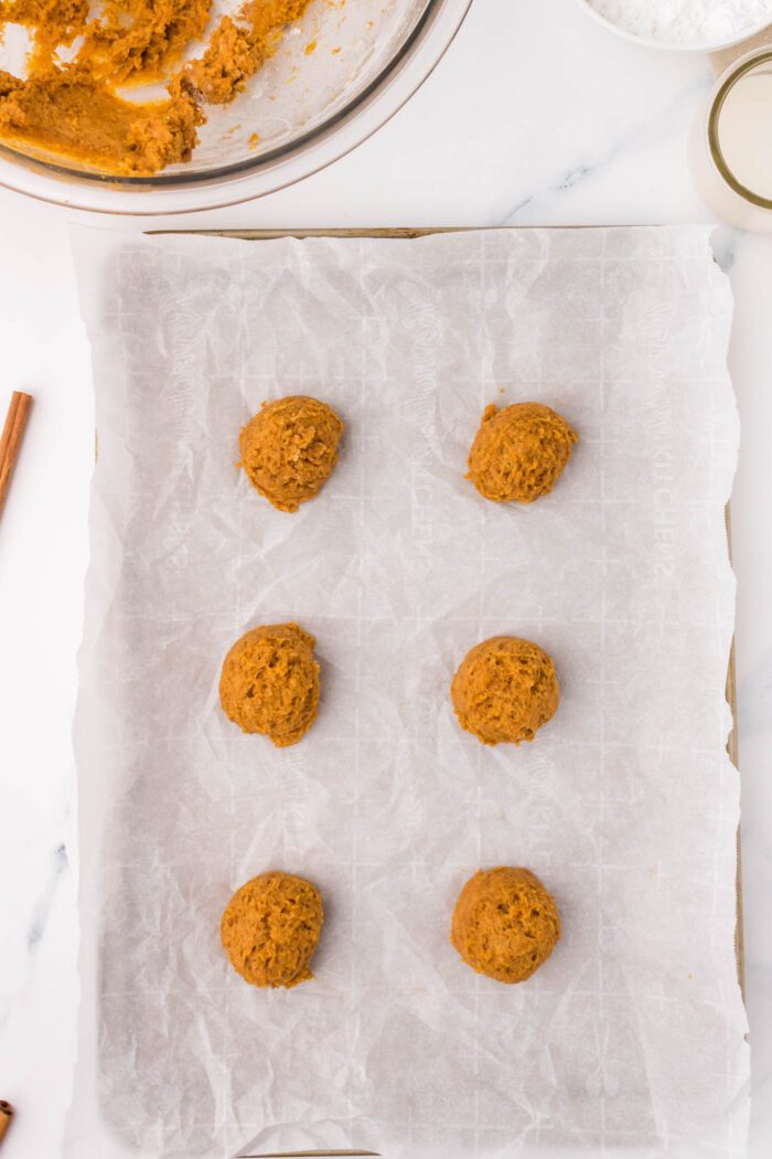 6 unbaked scoops of pumpkin cookie dough on a parchment paper-lined baking tray.