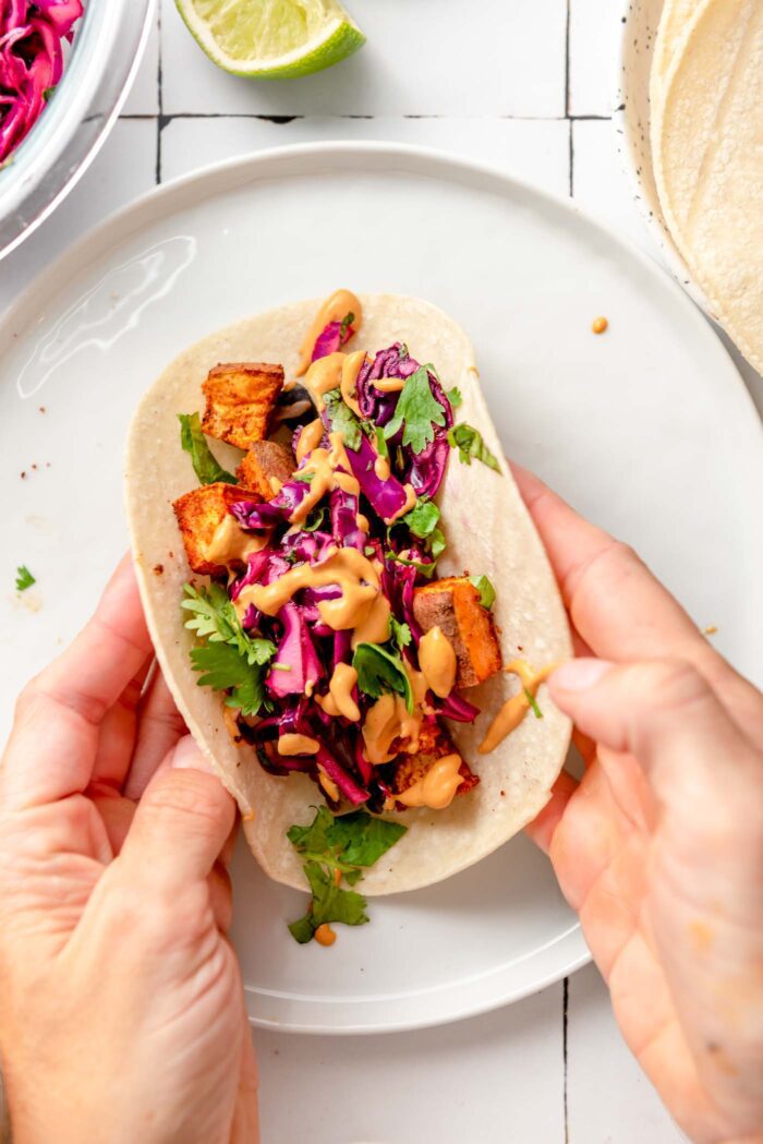Two hands picked up a sweet potato black bean taco with cabbage slaw, cilantro and queso sauce from a plate.