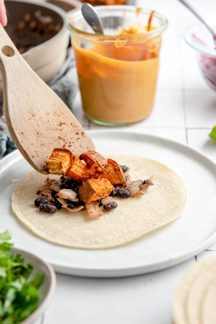 A corn tortilla on a plate. The tortilla is topped with an onion and black bean mixture with a wooden spoon scooping some spiced sweet potato cubes onto the tortilla. There's a glass jar of queso sauce in the background.