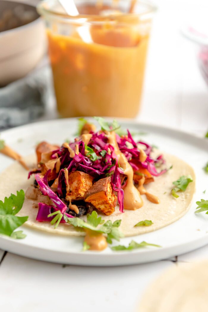 A colourful sweet potato and black bean taco topped with red cabbage slaw, cilantro and a creamy sauce on a plate. There's a jar of queso sauce with a spoon in it in the background.