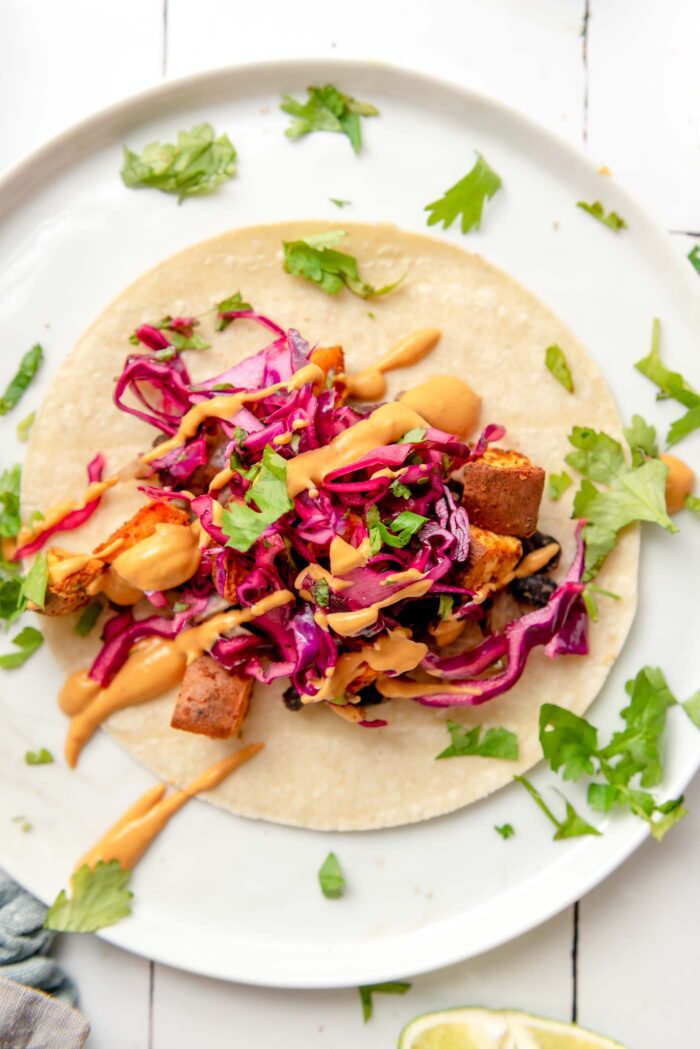 Overhead view of a colourful sweet potato and black bean taco topped with red cabbage slaw and a creamy sauce on a plate. Chopped fresh cilantro is sprinkled around the plate.
