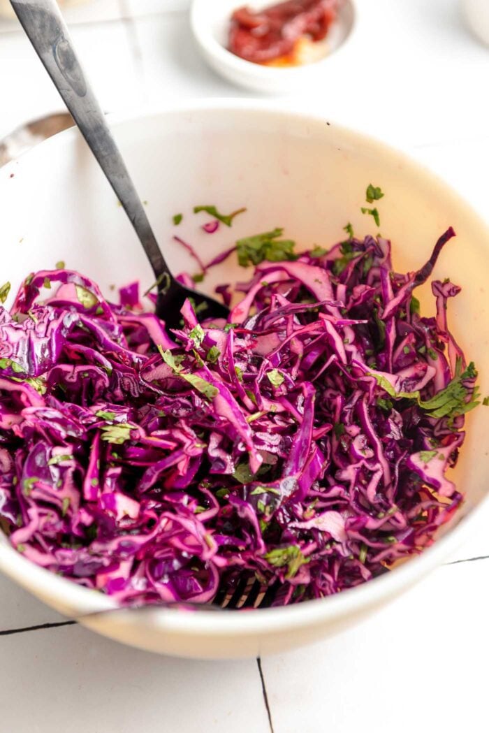 Red cabbage and cilantro slaw in a mixing bowl with a spoon and fork resting in the bowl.