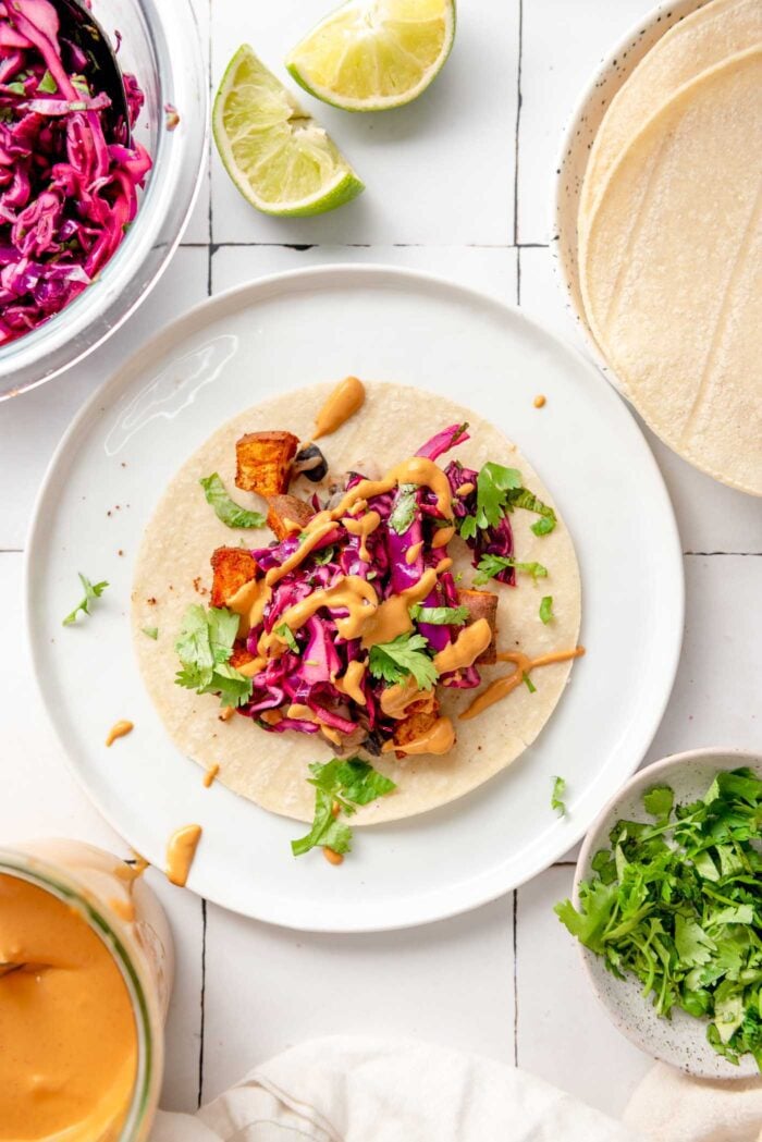Overhead view of a colourful sweet potato and black bean taco topped with red cabbage slaw and a creamy sauce on a plate. There is a plate of tortillas beside the plate and bowls of cilantro, cabbage slaw and the sauce.