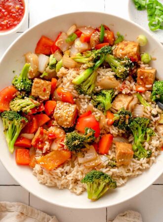 Close up overhead view of a bowl of a vegetable tempeh stir fry over rice. There are sesame seeds and chopped green onions scattered around the stir fry.