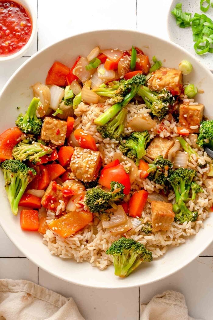Close up overhead view of a bowl of a vegetable tempeh stir fry over rice. There are sesame seeds and chopped green onions scattered around the stir fry.