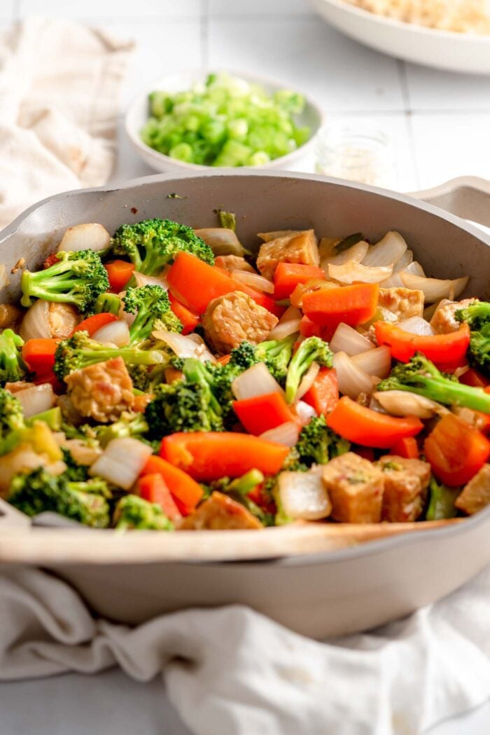 A vegetarian stir fry with tempeh, broccoli, bell pepper and onion in a skillet. There is a small dish of chopped green onions in the background.t