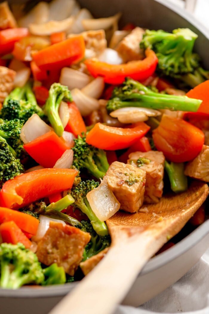 Close up of a wooden spoon resting in a skillet of stir fry with tempeh cubes and vegetables.