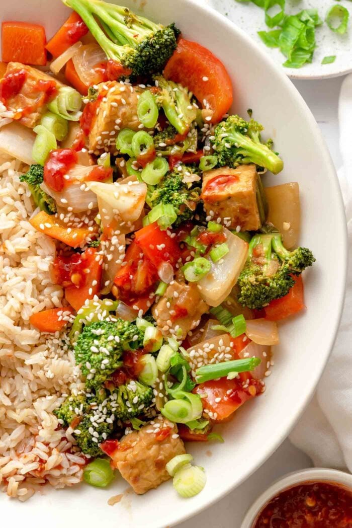Overhead view of a bowl of tempeh and vegetable stir fry with brown rice, sesame seeds and scallions. Only half of the bowl is in view showing mainly the stir fry portion with the brown rice just out of frame.