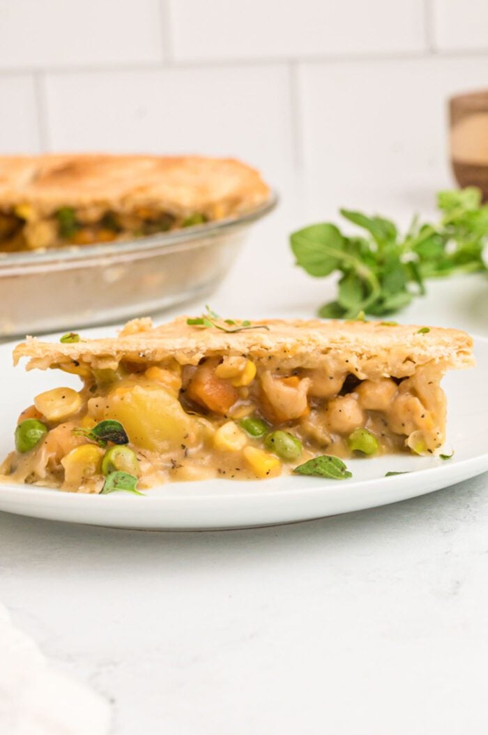 A slice of veggie pot pie with a potato, carrot, peas, corn and chickpea filling on a plate. The whole pie can be seen in the background.