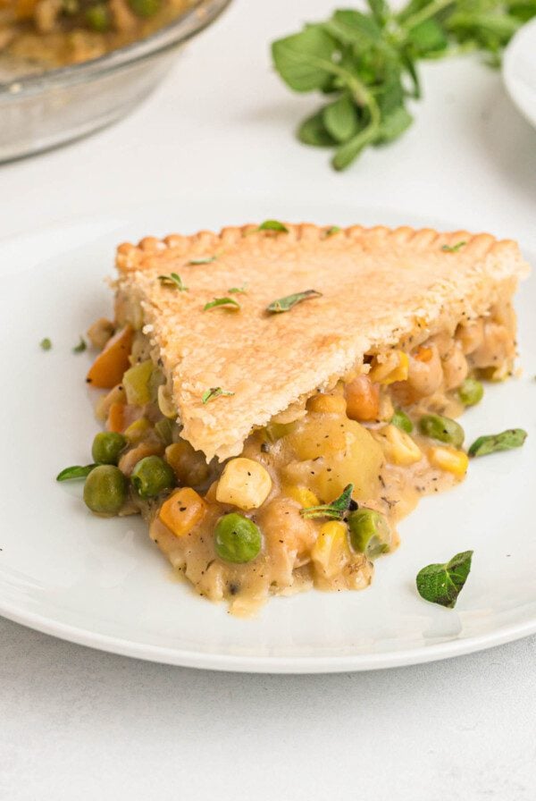 A slice of veggie pot pie with a potato, carrot, peas, corn and chickpea filling on a plate. The whole pie can be seen in the background.