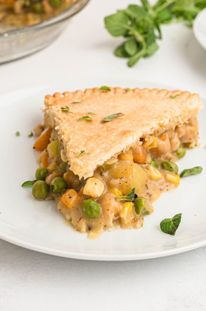 A slice of veggie pot pie with a potato, carrot, peas, corn and chickpea filling on a plate. The whole pie can be seen in the background.