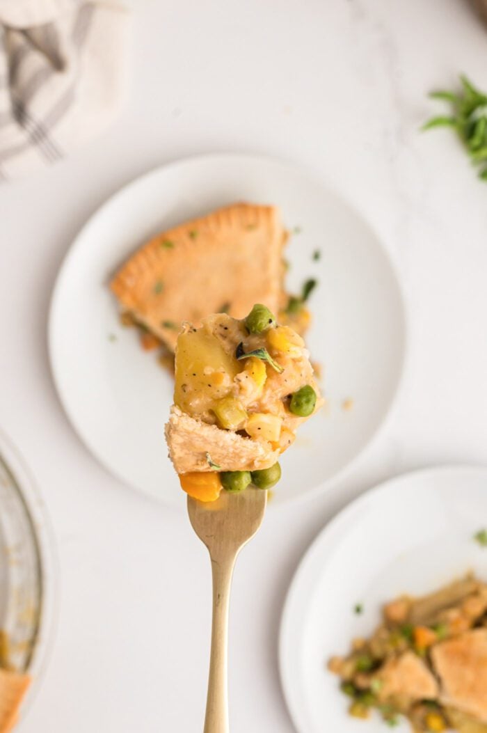 Fork with a bite-full of vegetable pot pie on it. The filling has peas, carrot and potato and there is a small piece of pie crust. The fork is held over a slice of pot pie on a plate.