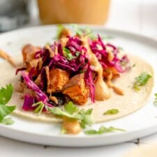 A colourful sweet potato and black bean taco topped with red cabbage slaw, cilantro and a creamy sauce on a plate. There's a jar of queso sauce with a spoon in it in the background.