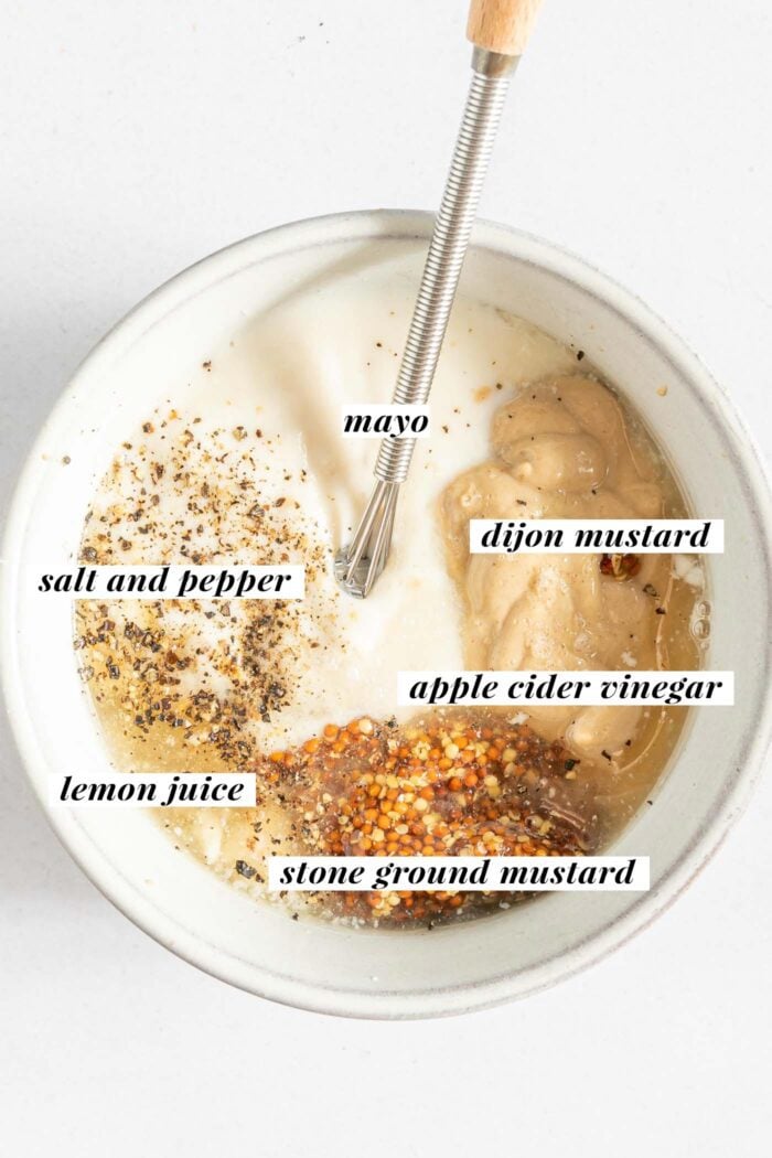 Mustard, mayonnaise, maple syrup, lemon juice, vinegar, salt and pepper in a small dish. A small whisk rests in the dish. Each ingredient is labelled with text.