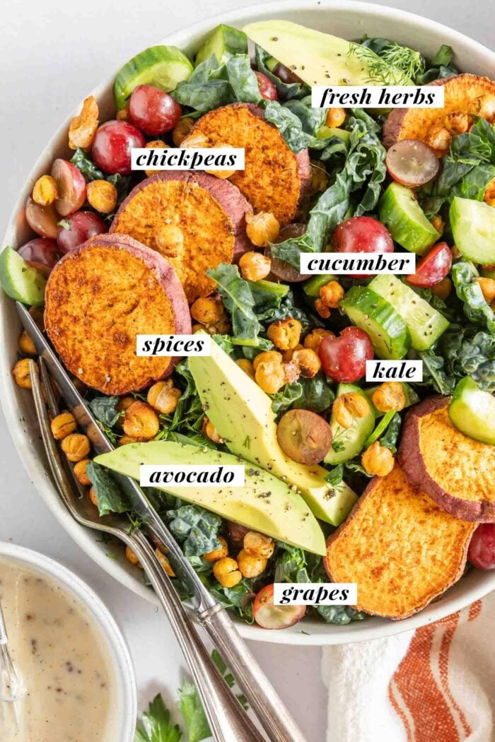 Overhead view of a avocado Tuscan kale salad with sweet potato, crispy chickpeas, grapes and avocado slices. A knife and fork rest in the bowl and there is a bowl of creamy dressing beside it. Each ingredient is labelled with text.
