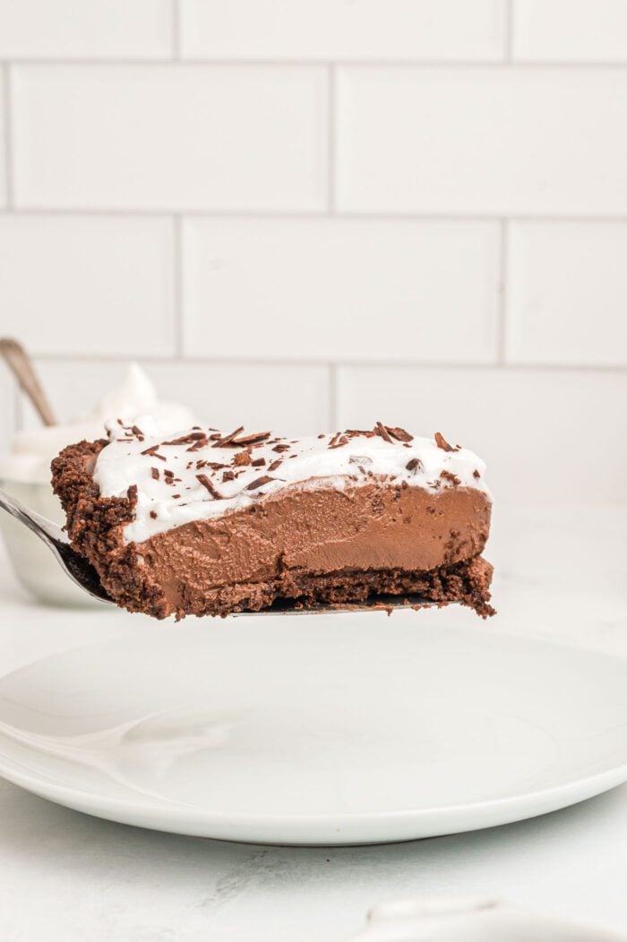 A slice of vegan chocolate pie with a chocolate crust topped with whipped cream on a spatula held over a plate.
