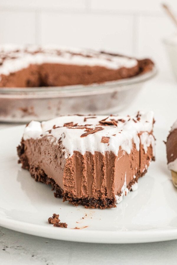 A slice of chocolate pie with chocolate crust topped with whipped cream and chocolate shavings on a small plate. The slice has a bite taken from it and you can see the marks a fork made when it sliced through the pie.