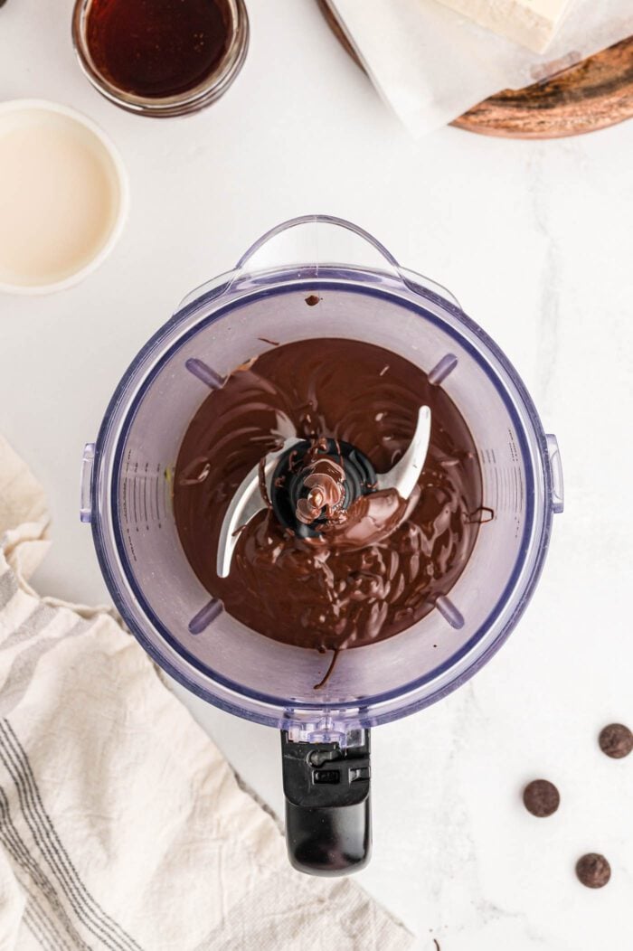 Melted chocolate in a food processor container with a few chocolate chips sprinkled around beside the container.