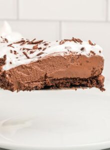 A slice of vegan chocolate pie with a chocolate crust topped with whipped cream on a spatula held over a plate against a white subway tile background.