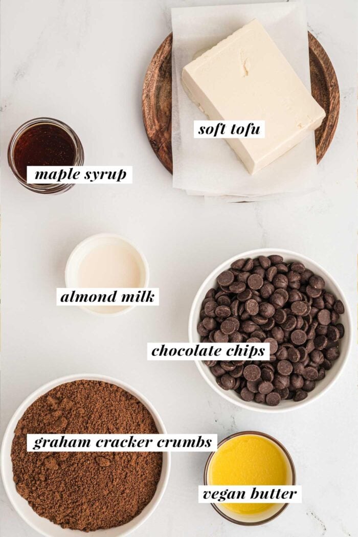 Graham cracker crumbs, melted butter, chocolate chips, almond milk, tofu and maple syrup in small bowls. Each is labelled with text describing the ingredient.