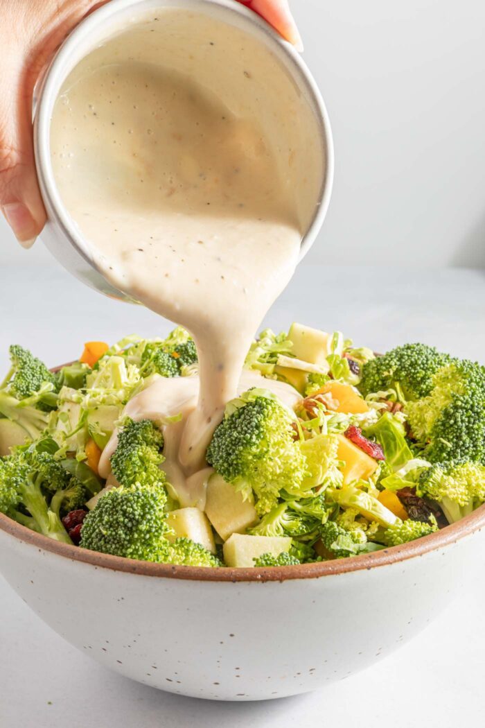 Creamy mayonnaise dressing being poured over a broccoli apple salad in a salad bowl.