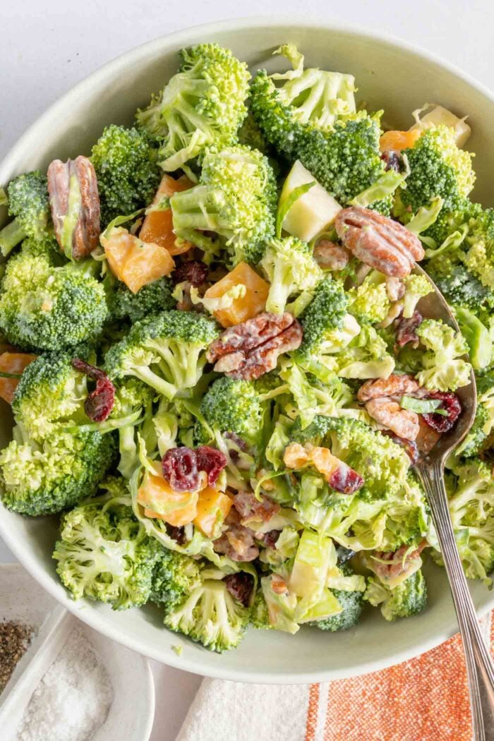 Overhead view of a bowl of broccoli apple salad with pecans, cranberries and brussels sprouts. There is a spoon resting in the bowl.