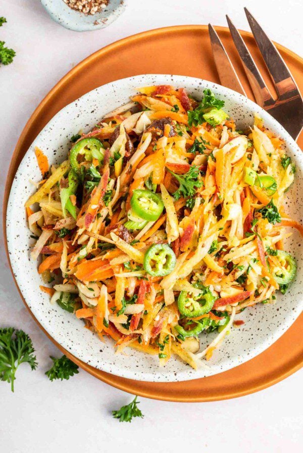 Overhead view of a carrot slaw with green onions, almonds, serrano peppers and parsley in a ceramic serving bowl. There's a large fork resting beside the bowl.