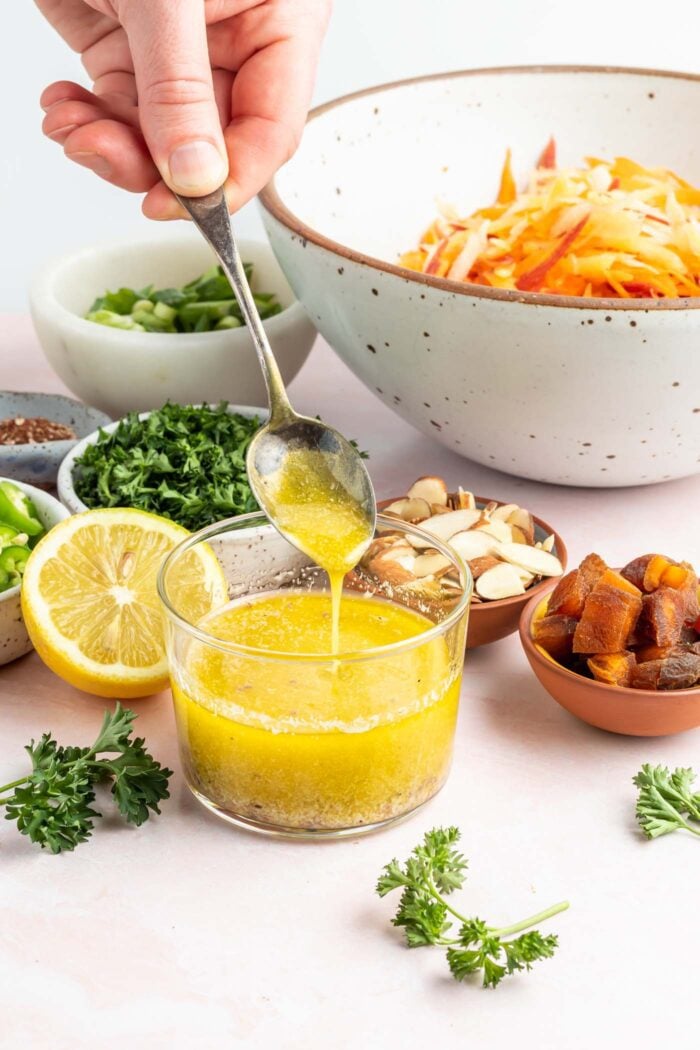 Hand using a spoon in a small bowl of lemon vinaigrette. There is a bowl of grated carrot behind the bowl of dressing.