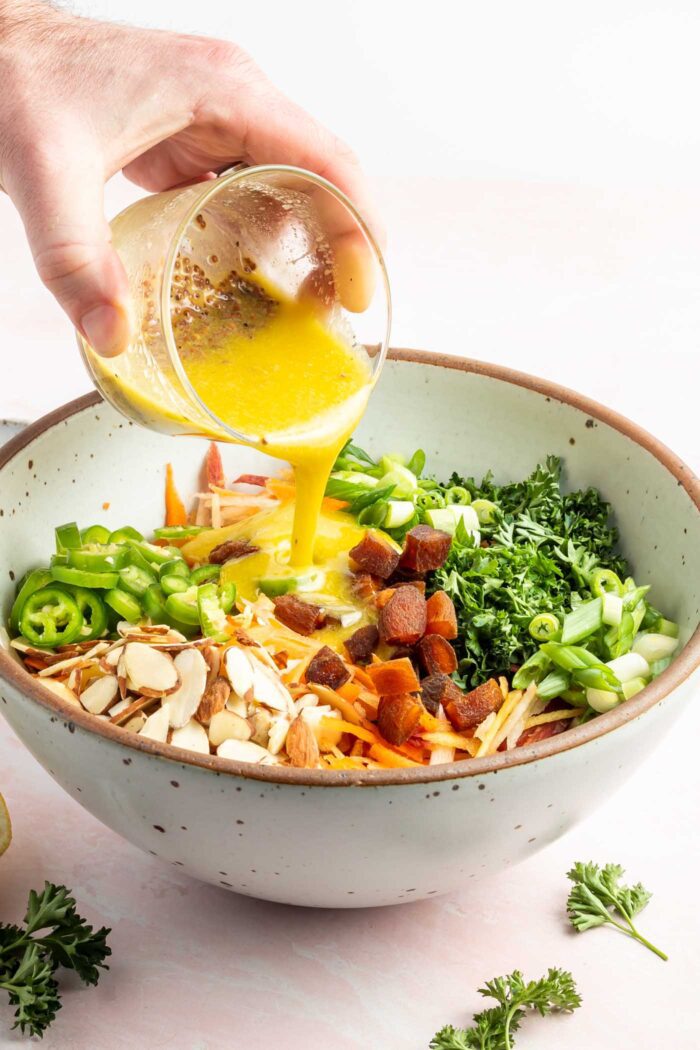Pouring a lemon vinaigrette from a small glass container over a grated carrot, green onion, almond and parsley in a ceramic salad bowl.