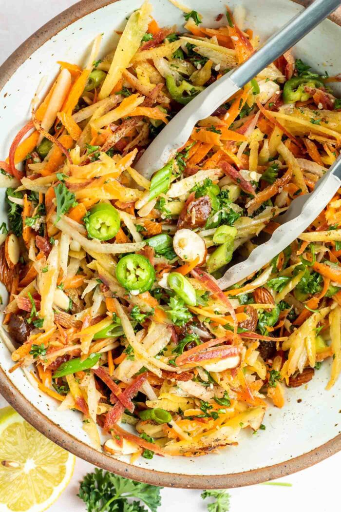 Overhead close up shot of a spicy carrot slaw with green onions, almonds, serrano peppers and parsley in a bowl. A pair of tongs rests in the bowl and there's a slice of lemon beside it.