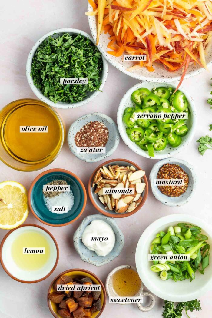 All the ingredients gathered in bowls for making a spicy carrot slaw recipe with mustard, almonds, lemon, green onion and apricot. Each ingredient is labelled with text describing the ingredient.