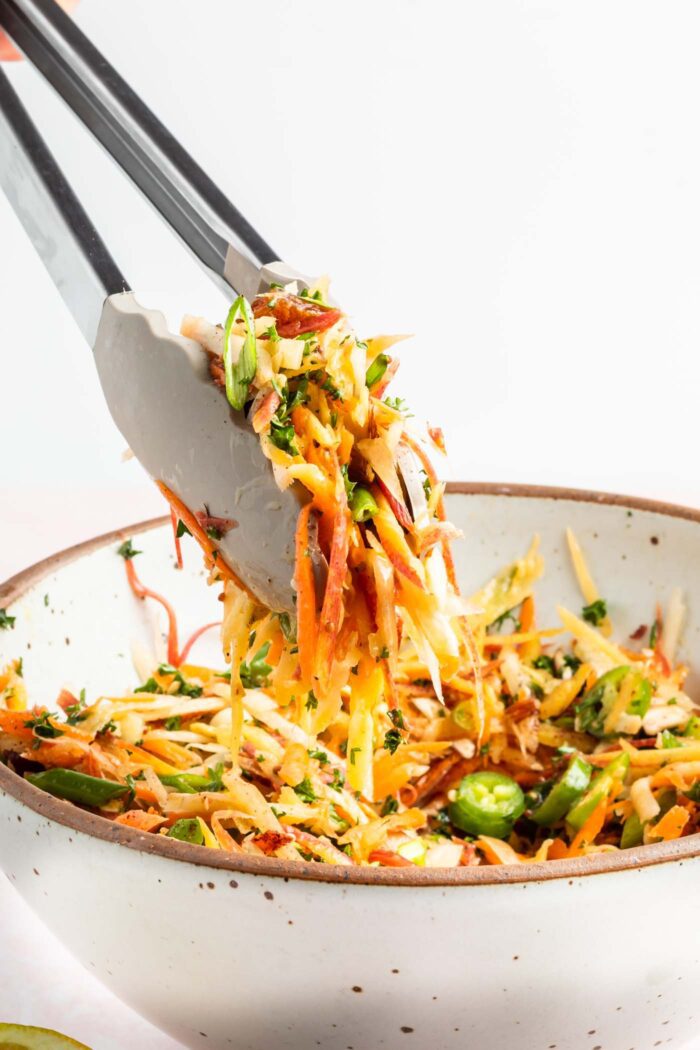 Tongs scooping spicy carrot slaw with serrano peppers, green onion and parsley from a serving bowl.