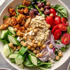 Overhead view of a colourful healthy bowl with spinach, hummus, tomato, red onion, cucumber and olives.