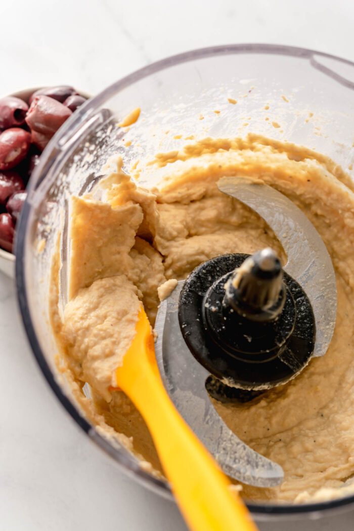Cremiger gemischter Hummus in einer Küchenmaschine mit einem kleinen Pfannenwender.  Neben der Küchenmaschine steht eine Schüssel mit Oliven.