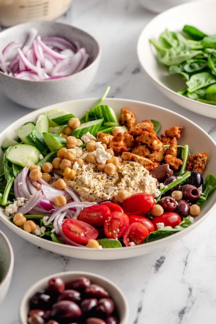 Healthy looking bowl with spinach, tofu, hummus, red onion, tomato, olives, cucumber and chickpeas. There are smaller bowls of sliced red onion, spinach and olives beside the bowl.