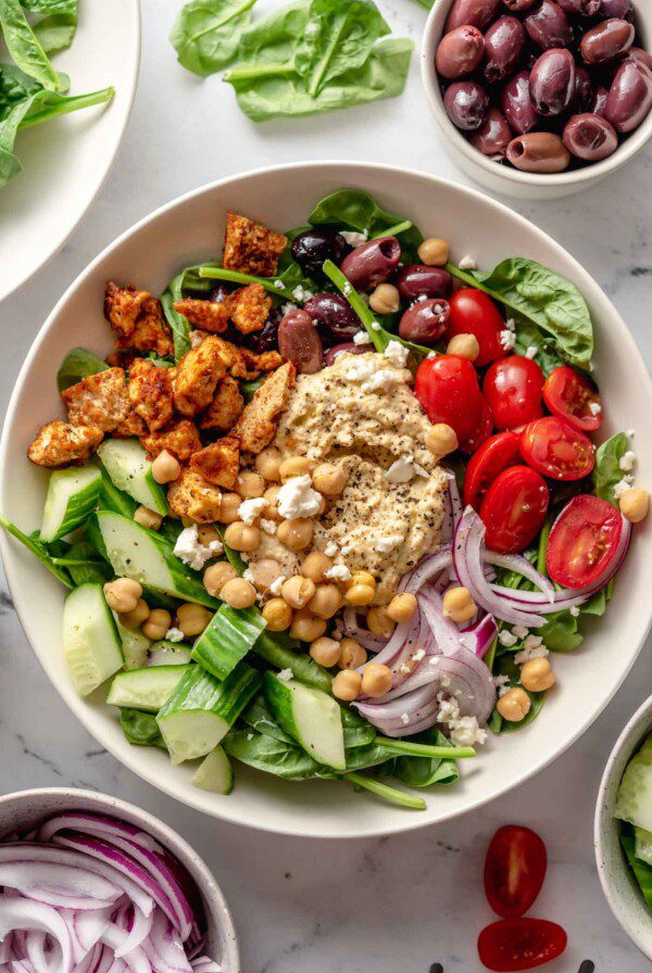 Overhead view of a colourful healthy bowl with spinach, hummus, tomato, red onion, cucumber and olives.