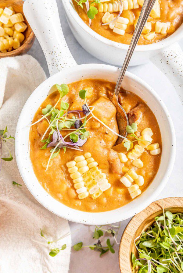 Overhead view of a handled bowl summer corn chowder soup topped with micro-greens and kernels of fresh corn. A spoon rests in the bowl and two additional bowls of soup are just out of frame.