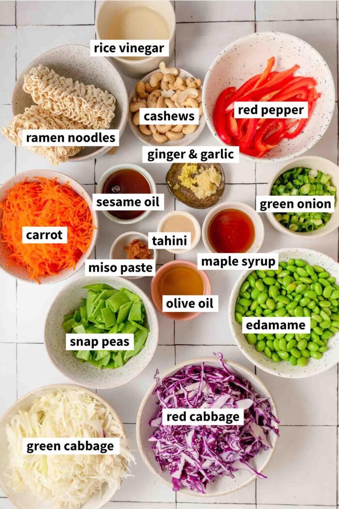 Ingredients for a ramen noodle salad with cabbage and other vegetables gathered in bowls. Each ingredient is labelled with text.