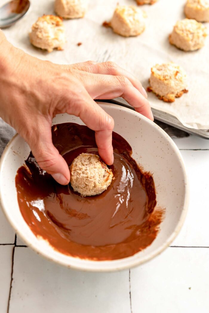 Hand dipping an almond macaroon into a bowl of melted chocolate. There is a baking tray of macaroons in the background.